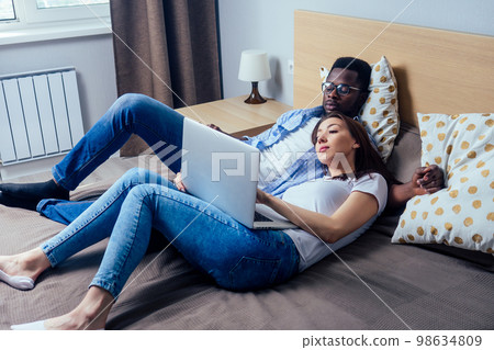 international couple smiling looking at laptop while resting on bed in bedroom together at home 98634809