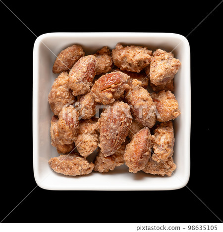 Candied almonds in a white square bowl. Homemade, in a special way cooked almonds, whole nuts coated in crunchy sugar. Sold at Christmas markets. Close-up, from above, isolated over black, food photo. 98635105