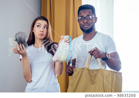 multinational couple choosing textile or polyester bag.african american man and asian woman save planet standing at living room multinational couple choosing textile or polyester bag.african american man and asian woman save planet standing at living room 98635208
