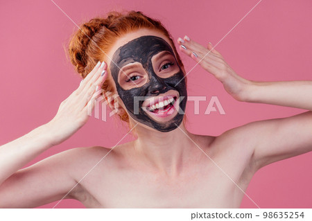 redhaired ginger teenager girl with black clay mask on her pretty face on pink studio background 98635254