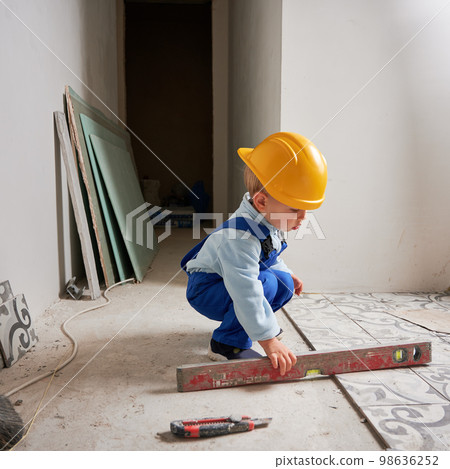 Child construction worker holding spirit level instrument. Kid in safety helmet and work overalls playing in apartment under renovation. Child construction worker holding spirit level instrument. Kid in safety helmet and work overalls playing in apartment under renovation. 98636252