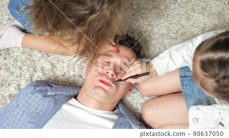 Girl siblings paint obedient father face with bright markers Girl siblings paint obedient father face with bright markers 98637050