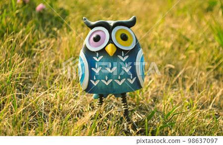 A toy of a surprised blue-colored owl with multi-colored eyes close-up stands in the middle of the grass in the light of the evening sun 98637097