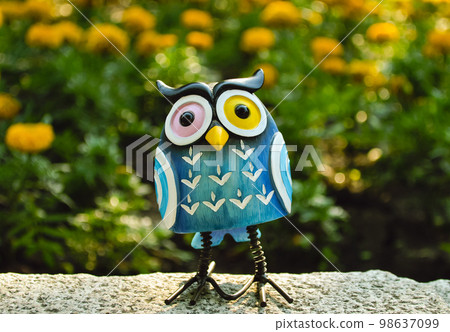 A toy of a surprised blue-colored owl with multi-colored eyes close-up stands on a stone against a background of yellow flowers in bokeh 98637099