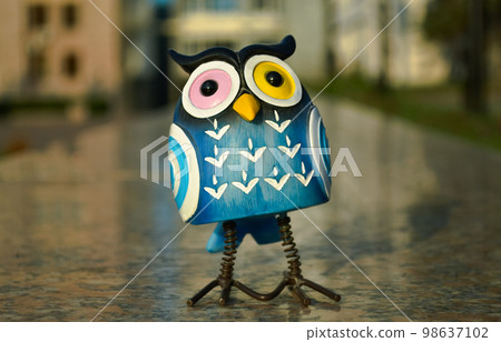 On polished granite, a surprised blue owl with multi-colored eyes stands close-up against the background of buildings in bokeh. Toy illuminated by evening sunlight 98637102