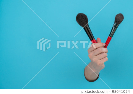 A woman is holding two large brushes for cosmetics. Woman's hand sticking out of blue paper background.  98641034