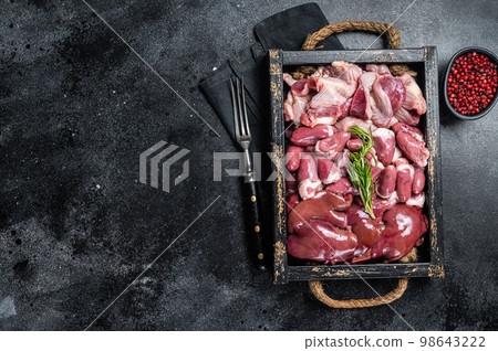 Uncooked Bird offals, raw chicken giblets, gizzards, stomachs, liver and hearts in a wooden tray. Black background. Top view. Copy space 98643222
