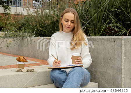 Young student study with notebook in park. Drinking coffee and eat croissants. Writing gratitude journal self reflection self discovery Young student study with notebook in park. Drinking coffee and eat croissants. Writing gratitude journal self reflection self discovery 98644325