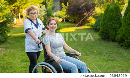 An elderly female nurse walks with a middle-aged woman in a wheelchair in the park.  98648919