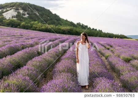 Close up portrait of happy young brunette woman in white dress on blooming fragrant lavender fields with endless rows. Warm sunset light. Bushes of lavender purple aromatic flowers on lavender fields. 98650763
