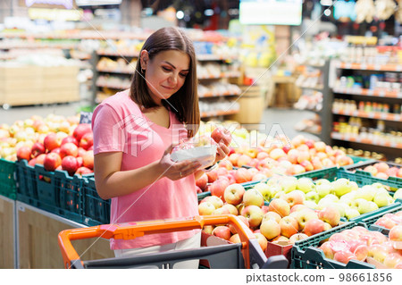 Young brunette woman picks chooses fruits on the counter in supermarket. Female housewife shopping in market standing near vegetable department store with a grocery cart. Young brunette woman picks chooses fruits on the counter in supermarket. Female housewife shopping in market standing near vegetable department store with a grocery cart. 98661856