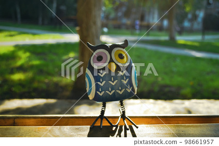 A close-up toy of a surprised blue owl with multi-colored eyes stands on a piece of wood in the light of the evening sun. The photo was taken in front of the sun 98661957