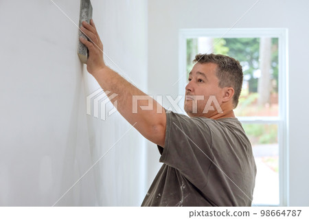 Worker sanding drywall mud in room during renovation of house with use sand trowel 98664787