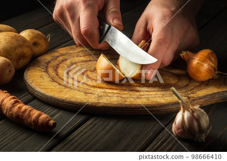 Slicing onions on a cutting board with a knife by the hands of a cook. Working environment in kitchen while preparing lunch chef Slicing onions on a cutting board with a knife by the hands of a cook. Working environment in kitchen while preparing lunch chef 98666410