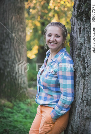 blonde in orange trousers and a plaid shirt smiling in the forest  98667578