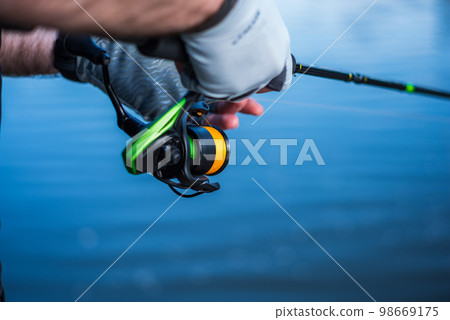 The fisherman holds a spinning rod in his hands. Angler with spinning rod and fishing reel. Hands and spinning on the background of water. 98669175