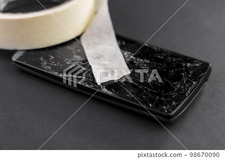 Broken smartphone with crushed touch screen display repaired with adhesive paper tape. Service maintenance and insurance of electronic device concept Broken smartphone with crushed touch screen display repaired with adhesive paper tape. Service maintenance and insurance of electronic device concept 98670090