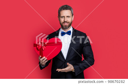 tuxedo man presenting valentine day proposal present isolated on red. man with valentine day gift tuxedo man presenting valentine day proposal present isolated on red. man with valentine day gift 98670282