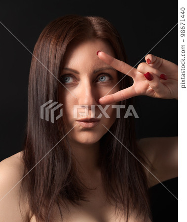 Studio portrait of 40-year-old woman showing victory or peace gesture with fingers near gray eyes. Caucasian ethnicity brunette with long hair and outstretched lips looking away on black background. Studio portrait of 40-year-old woman showing victory or peace gesture with fingers near gray eyes. Caucasian ethnicity brunette with long hair and outstretched lips looking away on black background. 98671440