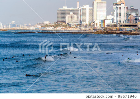 Several surfers  in distance ride the wave - other prepare to next waves. In distance hotrels of Tel Aviv and harbor of sailboats 98671946
