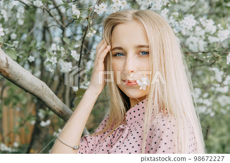 Blonde girl on a spring walk in the garden with cherry blossoms. Female portrait, close-up. A girl in a pink polka dot dress. Blonde girl on a spring walk in the garden with cherry blossoms. Female portrait, close-up. A girl in a pink polka dot dress. 98672227