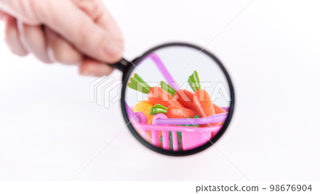 View through magnifying glass on plastic vegetables in shopping basket against white background. Nutrition value, GMO concept, plastic food, allergy, diet and food science, online shopping. 98676904