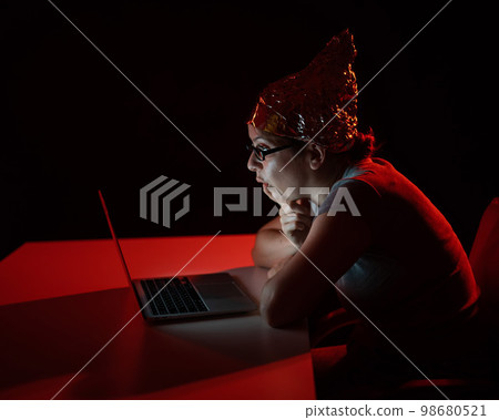 Paranoid caucasian woman typing on a computer wearing a tinfoil cap. Conspiracy theory. 98680521