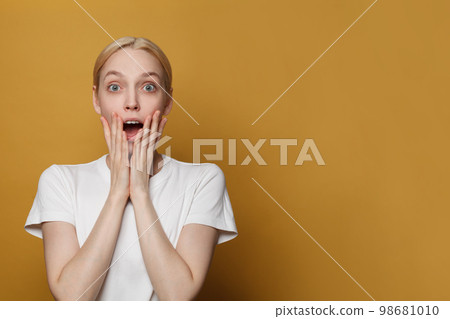 Surprised screaming blonde woman in white t-shirt looking at the camera over yellow studio wall 98681010