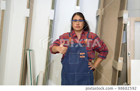 Asian middle aged woman with jean apron and protective glasses raise finger thumb up, stand infront plywood storage compartment. Carpenter worker work in a furniture factory. Asian middle aged woman with jean apron and protective glasses raise finger thumb up, stand infront plywood storage compartment. Carpenter worker work in a furniture factory. 98682592