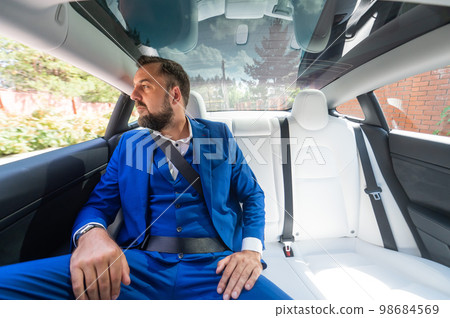 A caucasian man in a blue suit sits in the back seat of a car. Business class passenger. A caucasian man in a blue suit sits in the back seat of a car. Business class passenger. 98684569