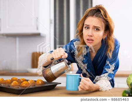 Sleepy woman in housecoat drinking morning coffee in kitchen Sleepy woman in housecoat drinking morning coffee in kitchen 98685463