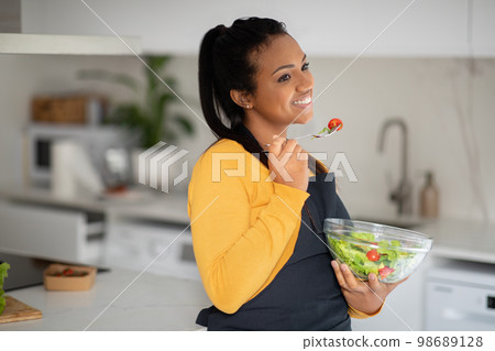 Cheerful pretty millennial african american lady in apron eat fresh vegetable salad in white kitchen interior 98689128