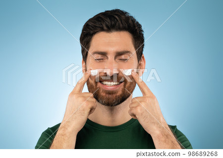 Happy bearded handsome man applying face cream on cheeks with closed eyes, posing over blue studio background 98689268