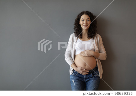 Portrait of young beautiful pregnant woman standing against grey wall indoors Portrait of young beautiful pregnant woman standing against grey wall indoors 98689355
