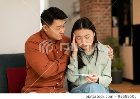 Infertility problem concept. Upset asian spouses holding negative pregnancy test, man calming wife, sitting on couch 98689503