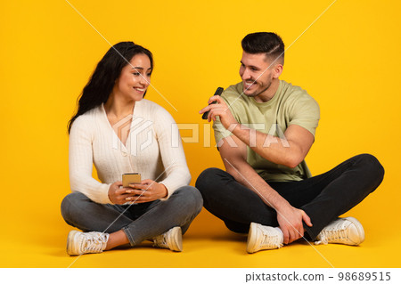 Cheerful young arab male in casual shows phone to wife, sit on floor isolated on yellow background, studio Cheerful young arab male in casual shows phone to wife, sit on floor isolated on yellow background, studio 98689515