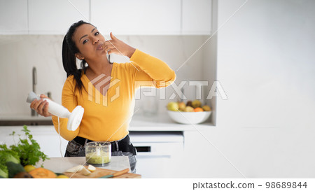 Cheerful millennial african american lady make smoothie with blender, licking finger at table with organic vegetables 98689844