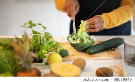 Millennial african american lady in apron cuts broccoli at table with vegetables in white kitchen interior, close up Millennial african american lady in apron cuts broccoli at table with vegetables in white kitchen interior, close up 98689849