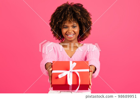 Cheerful pretty young black curly female in casual with braces gives red box with gift Cheerful pretty young black curly female in casual with braces gives red box with gift 98689951
