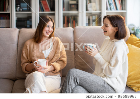 Laughing european millennial ladies in sweaters with coffee cups talk, sit on sofa, enjoy visit, free time 98689965