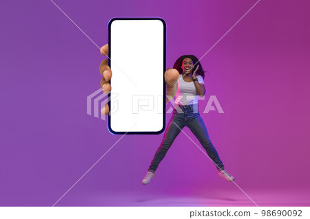 Black Excited Woman Jumping With Blank Smartphone In Hand Under Neon Light Black Excited Woman Jumping With Blank Smartphone In Hand Under Neon Light 98690092