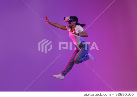 Excited Black Woman Wearing VR Glasses Jumping In Air In Neon Light Excited Black Woman Wearing VR Glasses Jumping In Air In Neon Light 98690100