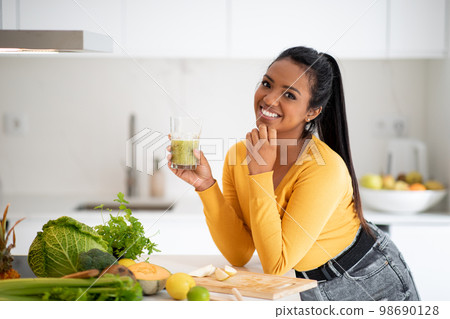 Smiling young african american female vegan at table with organic vegetables hold glass with fresh smoothie 98690128