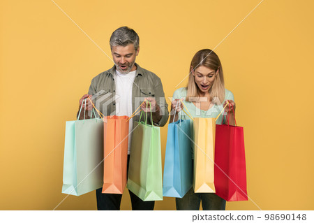 Shopaholics. Overjoyed middle aged caucasian couple holding shopping bags and looking inside, yellow background Shopaholics. Overjoyed middle aged caucasian couple holding shopping bags and looking inside, yellow background 98690148