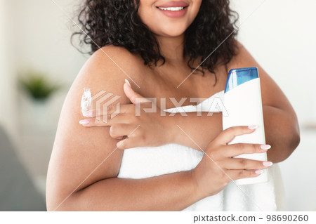 Young plus size woman showing lotion bottle, advertising cosmetics and applying product on her shoulder after bathing 98690260