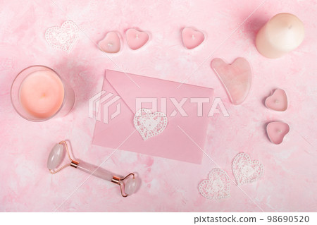 Flat lay concept for face spa skin treatment. Natural pink quartz stone Guasha scrape and roller massager for face, envelope, different candles on pink background. Gift certificate image. Top view. 98690520