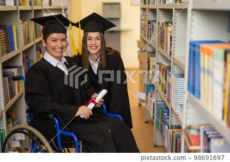 Happy young woman and woman in wheelchair in graduate gown with diploma in hands in library. Inclusive education. Happy young woman and woman in wheelchair in graduate gown with diploma in hands in library. Inclusive education. 98691697