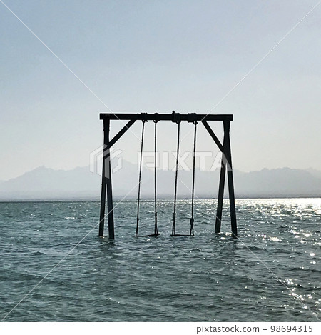 Wooden childrens swing stands in blue calm sea under open clean sky Wooden childrens swing stands in blue calm sea under open clean sky 98694315