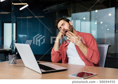 A tired young man in a red shirt sits at the desk in the office and yawns, covering his mouth with his hand. Works overtime, boring monotonous work, falls asleep. 98694450