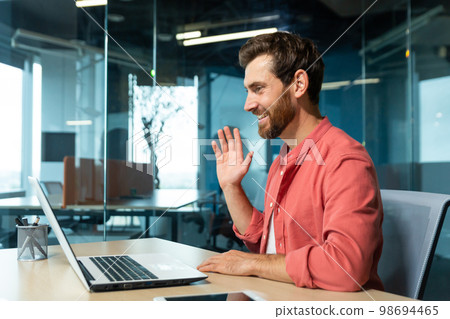 Portrait of a young man in a red shirt. Sitting in the office at the table, working on a laptop, communicating via video call, waving, smiling. Portrait of a young man in a red shirt. Sitting in the office at the table, working on a laptop, communicating via video call, waving, smiling. 98694465
